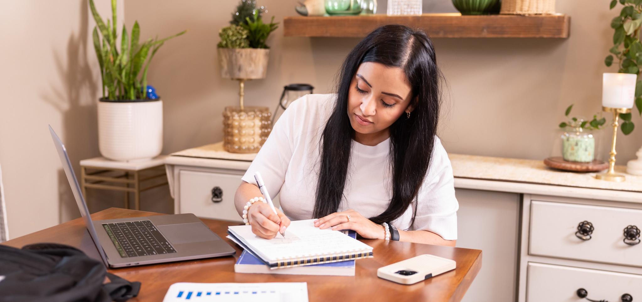 Woman writing in a notebook at a table with a laptop and smartphone, surrounded by plants and decor.