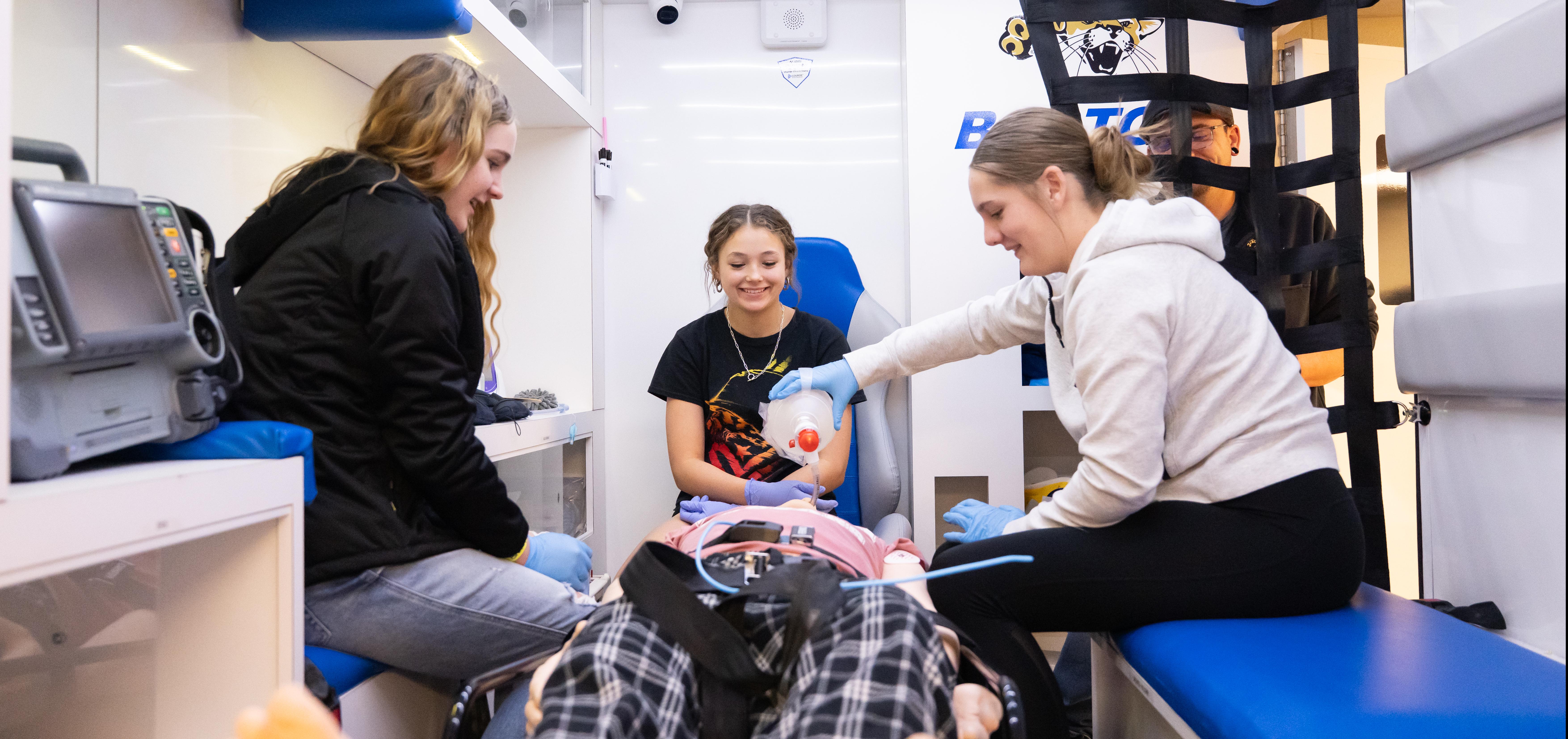 Three students sit in the ambulance simulator, smiling and practicing intubating a mannequin.